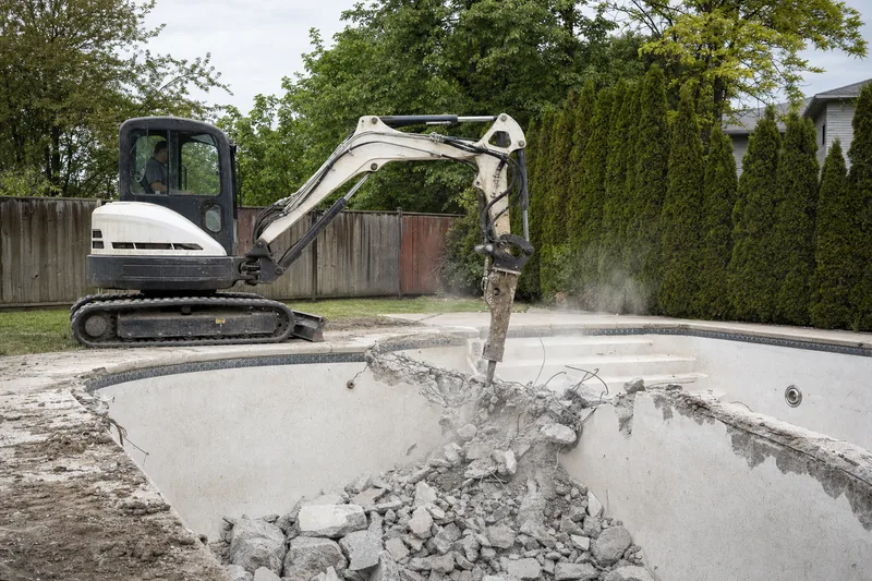 An excavator with a hydraulic hammer breaking the wall of an empty in-ground pool — pool removal, Geelong. Illustrative example.